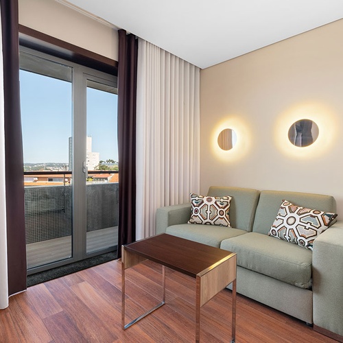 Uma sala de estar moderna apresenta um sofá verde com almofadas estampadas, uma mesa de centro, luminárias de parede circulares e uma porta de correr para a varanda, tudo sobre um piso de madeira.