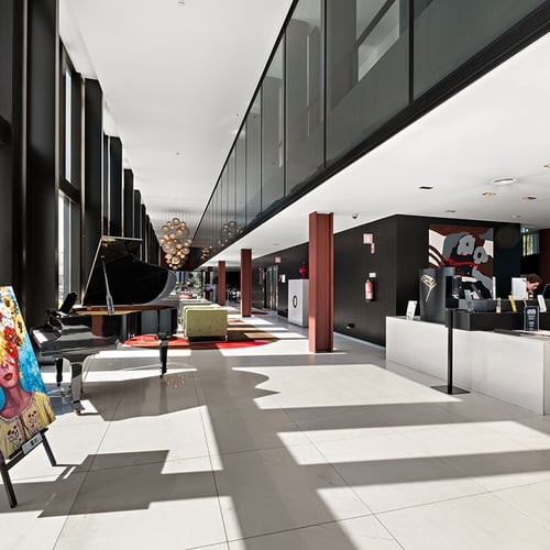 Uma ampla vista interior revela um lobby moderno e espaçoso, adornado com grandes janelas, um piano de cauda, uma vibrante pintura num cavalete e uma área de recepção.
