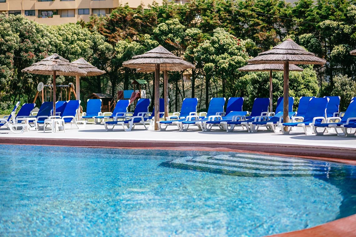 Espreguiçadeiras azuis e guarda-sóis de palha junto à piscina de um hotel. Vermar | Axis Hotéis & Golfe | Website Oficial