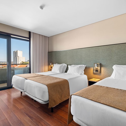 Este é um quarto de hotel com três camas individuais, chão de madeira, uma varanda que oferece vista para a cidade e um edifício alto ao longe.