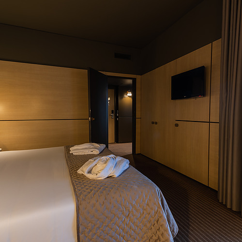 Quarto de hotel com cama de casal, roupões e porta aberta para outro cômodo. Axis: Corporate.