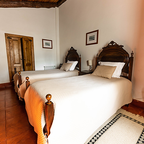 Camas gemelas con dosel de madera oscura y edredones blancos, baño con azulejos de terracota, vigas de madera en el techo. Axis: Corporate