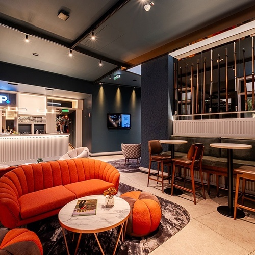 A stylish and modern hotel lounge or bar area features a vibrant orange sofa and ottoman, a sleek white bar with an 
