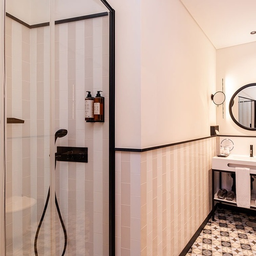 This modern bathroom features a glass-enclosed shower with vertical striped wall tiles, a vanity with a round illuminated mirror and black fixtures, and patterned geometric floor tiles.