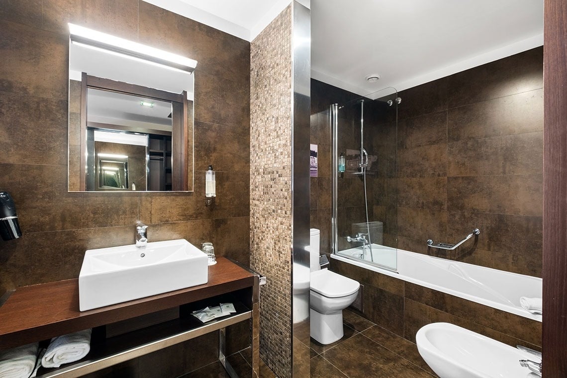 Um banheiro moderno com revestimento em azulejos marrons, equipado com pia, espelho, banheira com chuveiro, vaso sanitário e bidê.