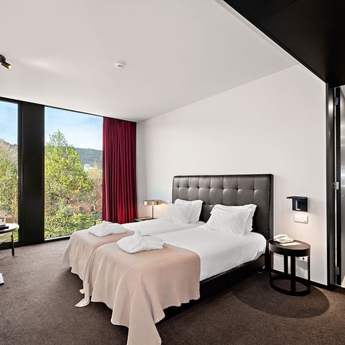 Quarto de hotel moderno com duas camas de solteiro, uma grande janela com vista para a natureza e um vislumbre da casa de banho.