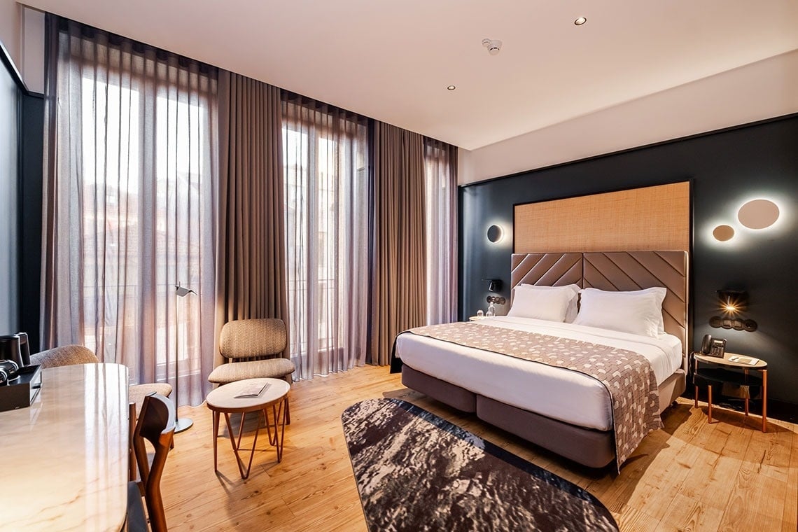 Um quarto de hotel moderno e elegante, com uma cama grande, parede de destaque escura, janelas amplas com cortinas e piso de madeira.