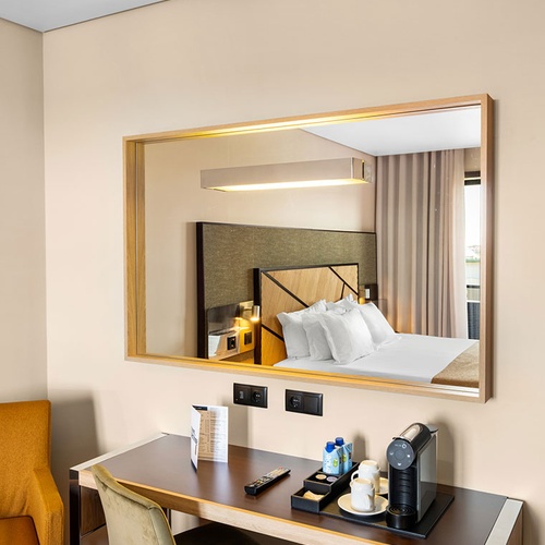 Um quarto de hotel moderno exibe uma secretária equipada com máquina de café e um grande espelho refletindo uma cama confortável.