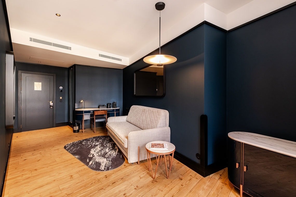 Um quarto moderno apresenta paredes azul-marinho, piso de madeira clara e móveis contemporâneos, incluindo um sofá bege, uma secretária e um aparador.