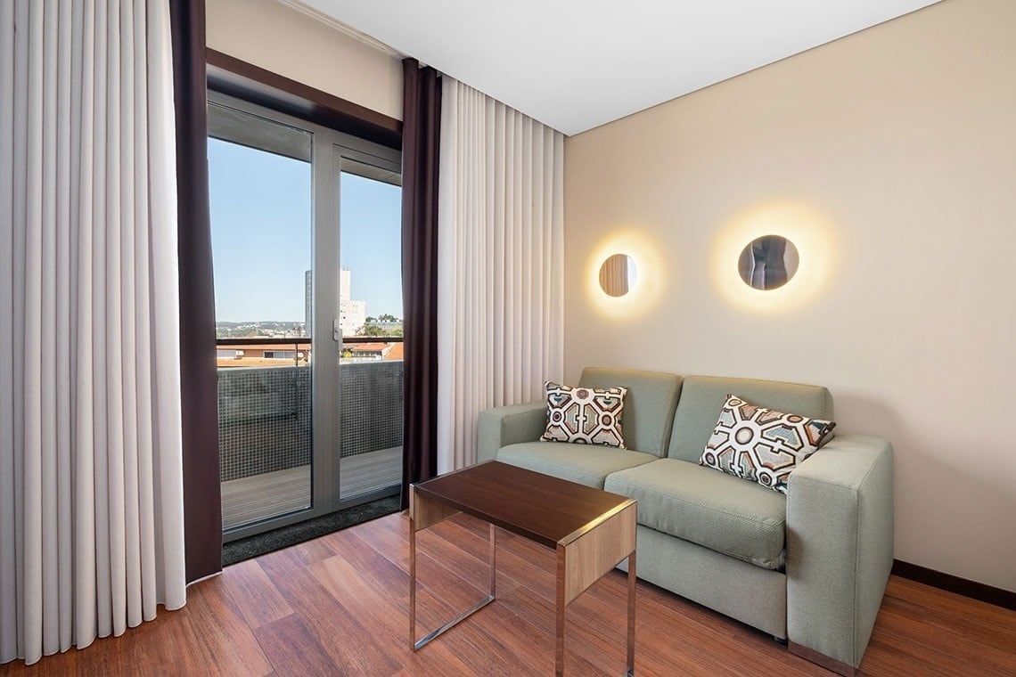 Uma sala de estar moderna apresenta um sofá verde com almofadas estampadas, uma mesa de centro de madeira e uma porta de vidro que se abre para uma varanda, complementada por arandelas circulares na parede e cortinas bicolores.