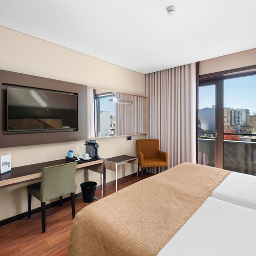 Um quarto de hotel moderno com cama, secretária com televisão e uma varanda com vista para o exterior.