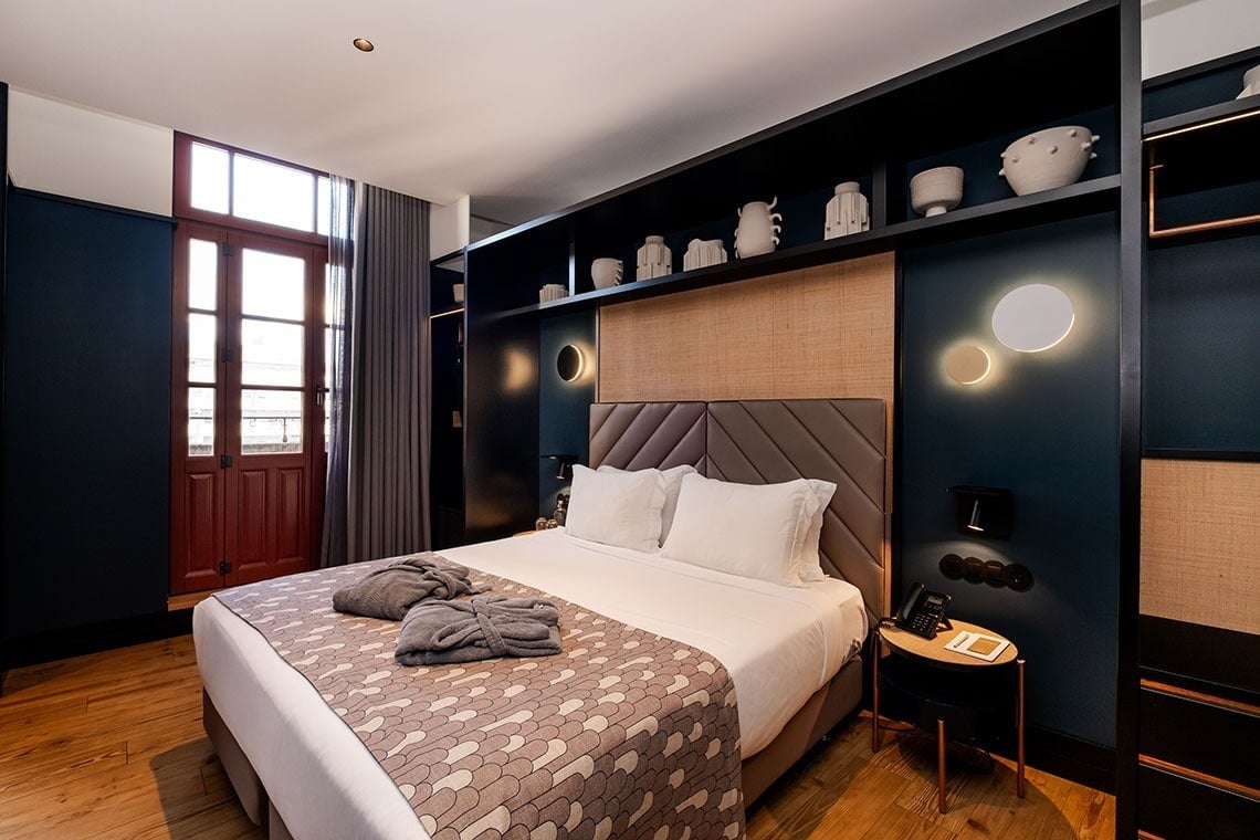Um quarto de hotel moderno e elegante, com uma cama grande, cabeceira acolchoada, estantes decorativas e uma porta de madeira que leva ao exterior.