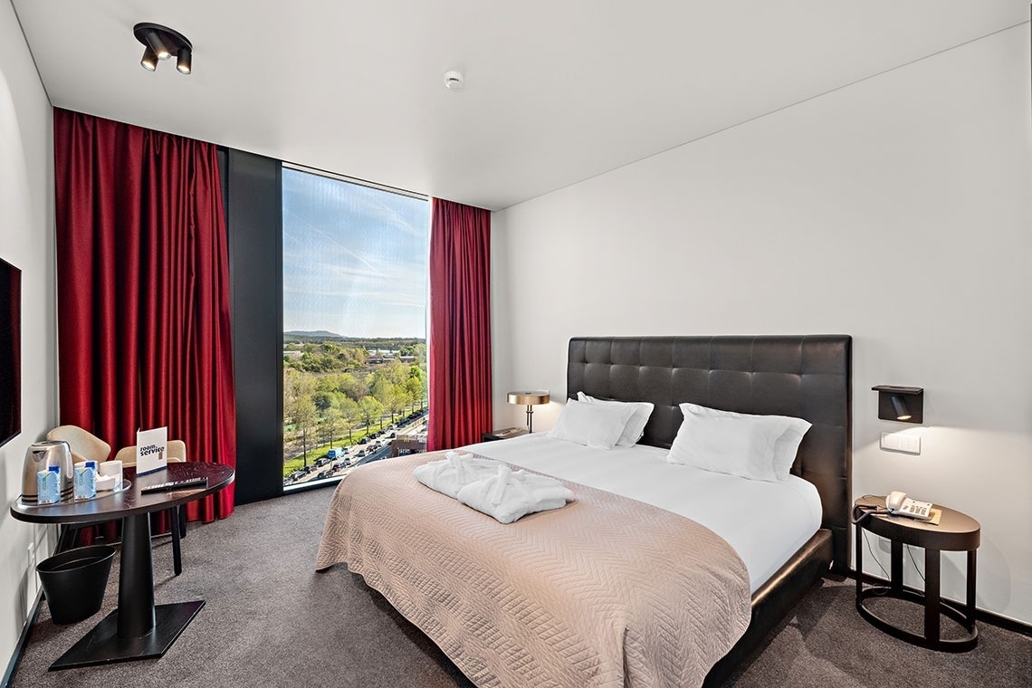 Une chambre d'hôtel moderne est aménagée avec un grand lit, une table de service sur la gauche, et une vaste fenêtre offrant une vue panoramique sur la ville et la verdure, encadrée par des rideaux rouges.