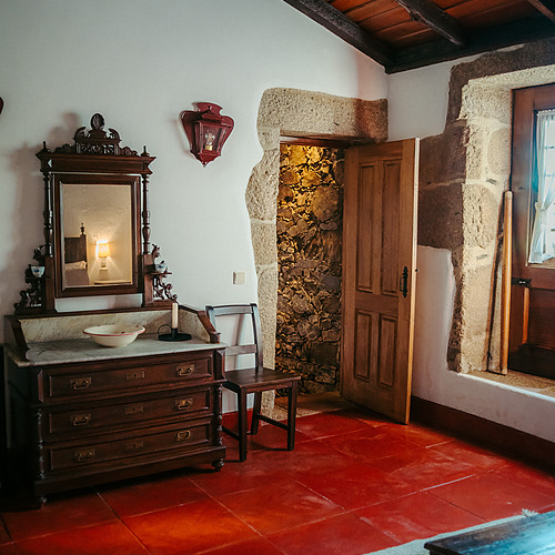 Habitación rústica con suelo rojo, tocador antiguo y puerta de piedra. Axis: Corporate