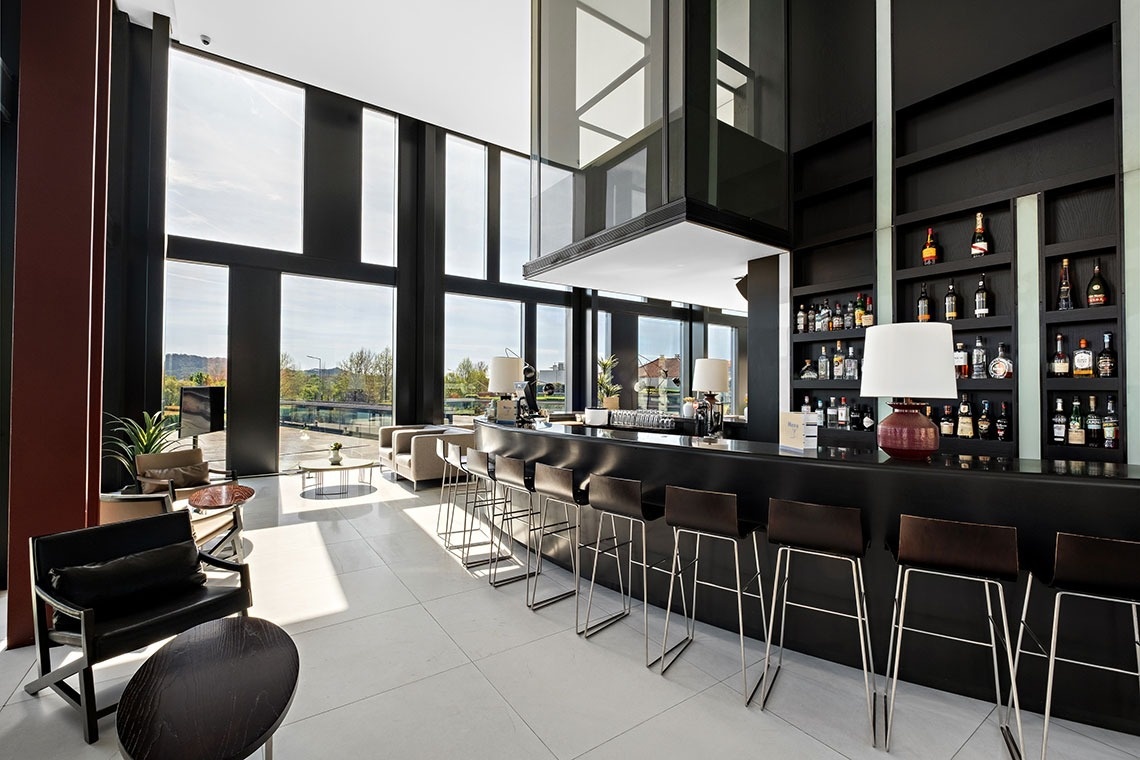 Un espace bar/lounge moderne et lumineux doté de grandes baies vitrées, d'un bar noir élégant et d'un ascenseur en verre.