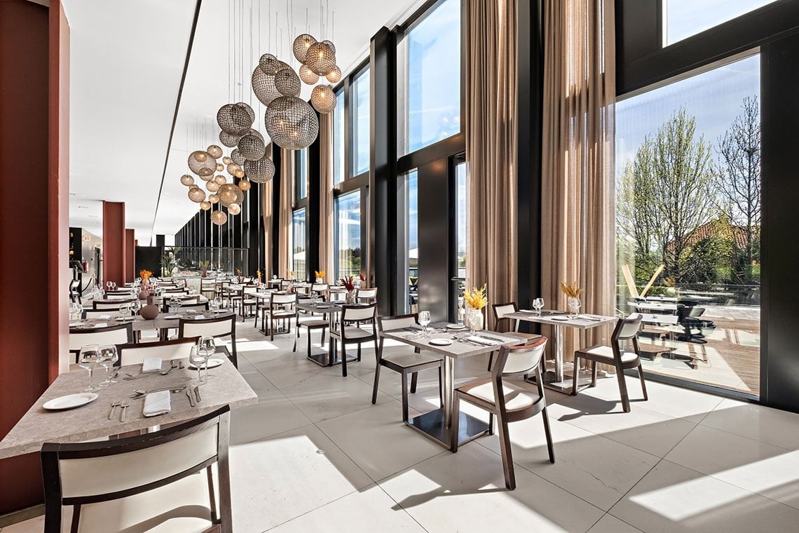 Une salle de restaurant moderne et lumineuse est caractérisée par de grandes baies vitrées, des luminaires sphériques originaux et de nombreuses tables dressées.