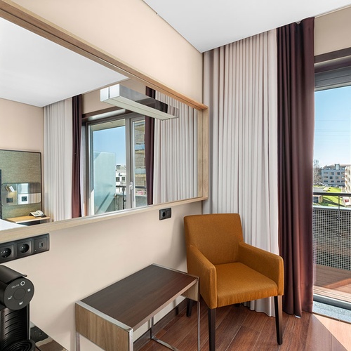 Um quarto moderno apresenta um espelho grande, uma cafeteira, uma poltrona laranja e uma porta de varanda com vista para edifícios e vegetação.