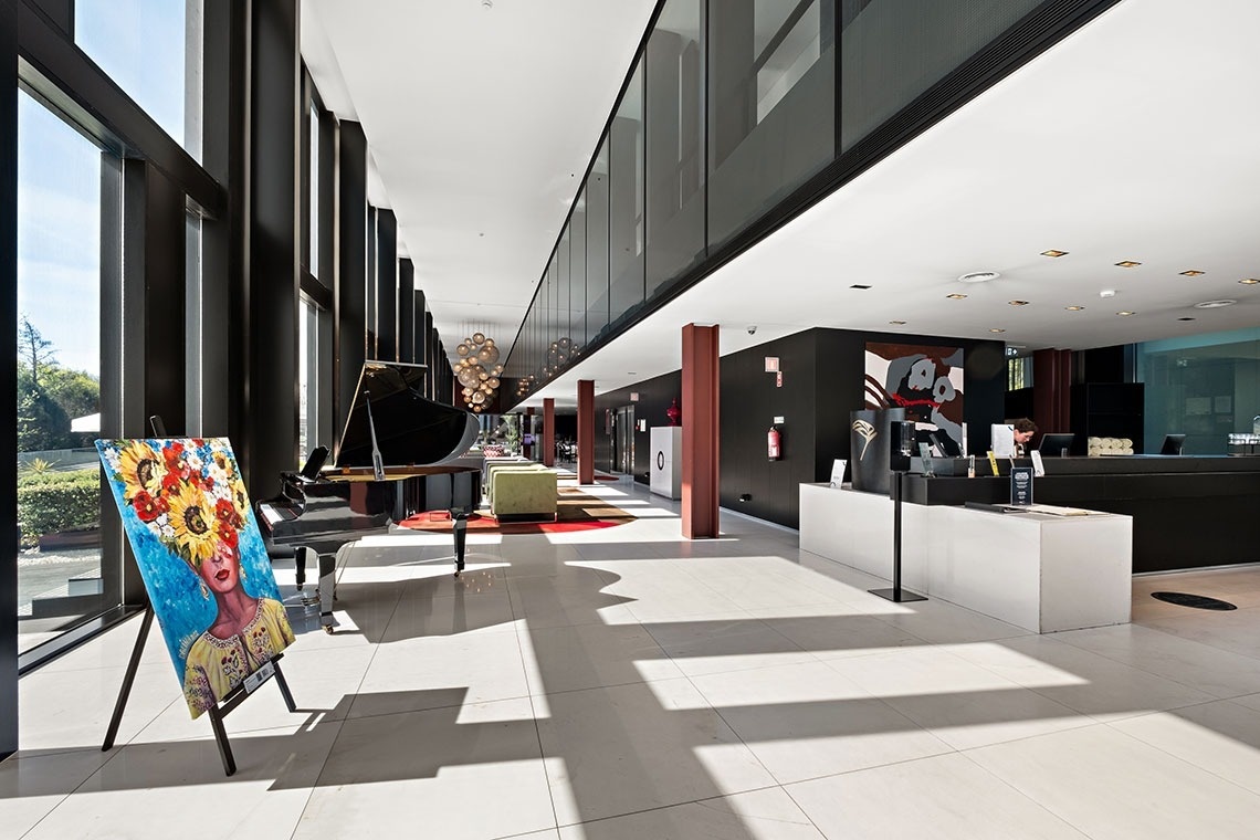 Une vue d'un hall moderne et lumineux avec de grandes baies vitrées, un piano à queue, un tableau coloré sur un chevalet, un comptoir de réception et un niveau supérieur avec une balustrade en verre.