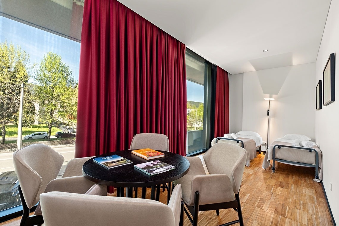 Cette chambre moderne et lumineuse est aménagée avec de grandes baies vitrées parées de rideaux rouges, un coin salon central doté d'une table ronde et de fauteuils, et deux lits individuels.