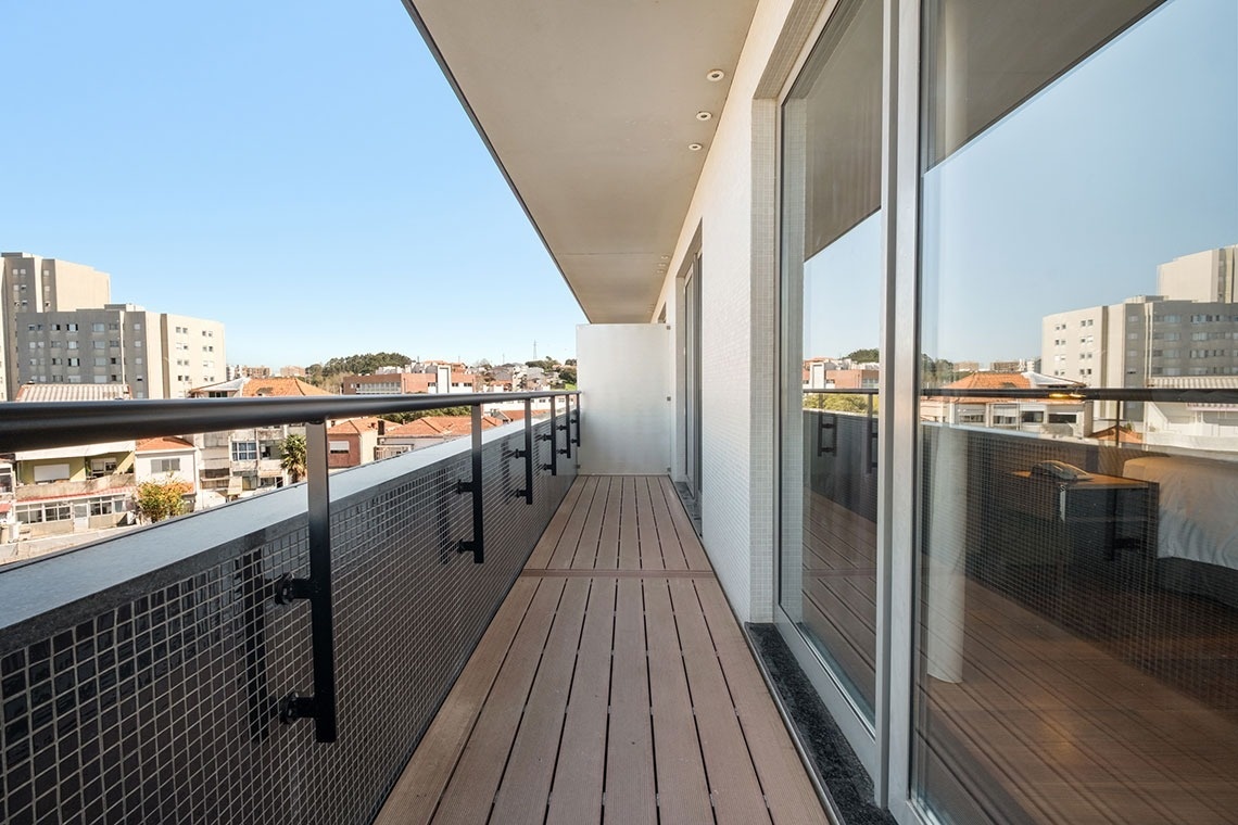 pt: Uma varanda moderna e comprida, com deck de madeira, guarda-corpo de metal sobre revestimento de pastilhas escuras, e portas de vidro que refletem o interior de um quarto, com uma vista urbana ao fundo.