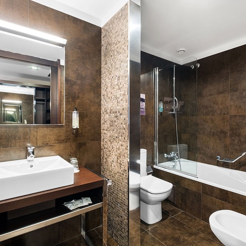 Este é um banheiro moderno com revestimentos em tons de marrom, destacando-se o lavatório, vaso sanitário, banheira com chuveiro e um bidê, além de um detalhe em mosaico.