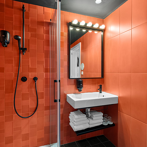 Baño moderno con ducha, lavabo, espejo iluminado y azulejos naranjas vibrantes. Ideal para unAxis: Corporate.