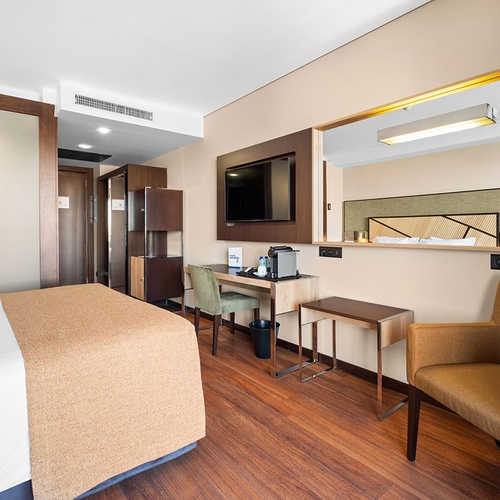 Um quarto de hotel moderno e luminoso, com uma cama arrumada, piso de madeira, secretária com TV e poltrona, apresentando um design elegante em tons neutros.