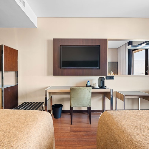 Um quarto de hotel moderno e bem iluminado, apresentando duas camas, uma televisão montada na parede sobre uma secretária com máquina de café, e um espelho que reflete a vista exterior.