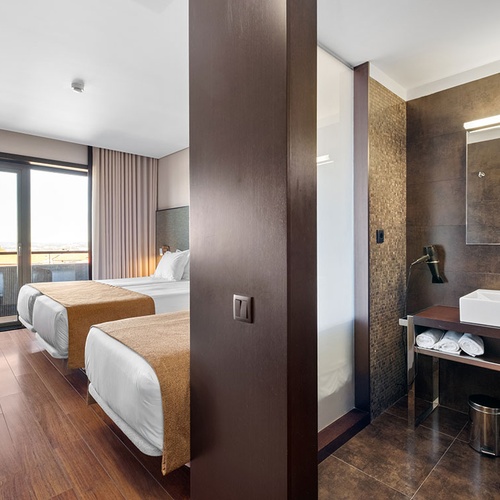 Um quarto de hotel moderno e funcional, com duas camas, uma sacada com vista e uma área de lavatório integrada.