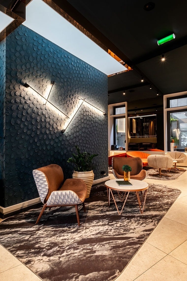 Uma área de lounge moderna e sofisticada é destacada por uma parede azul escura texturizada com iluminação linear, poltronas confortáveis em tons de terra e um tapete com padrão orgânico.