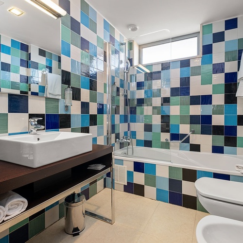 Uma casa de banho moderna apresenta paredes revestidas com azulejos quadriculados em tons de azul, verde e branco, equipada com uma pia retangular, banheira com chuveiro, vaso sanitário e bidê.