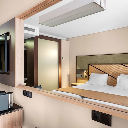 Um quarto de hotel moderno com cama de casal, cabeceira de madeira geométrica e televisão, em parte visível através de um espelho grande.