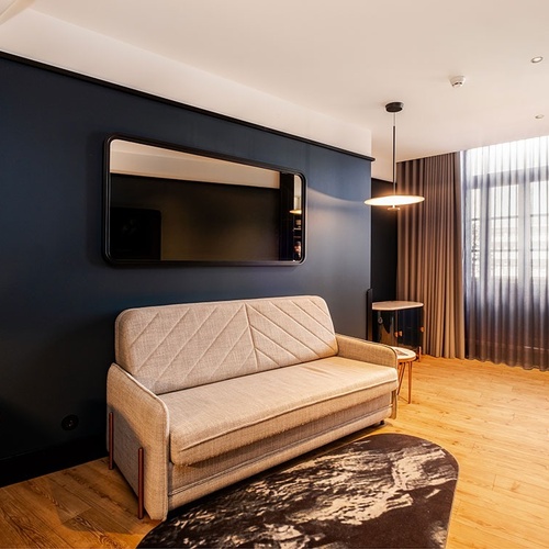 A modern living area features a light-colored sofa against a dark blue wall with a large rectangular mirror above it, a wooden floor, a dark patterned rug, and a window covered by sheer and opaque curtains.
