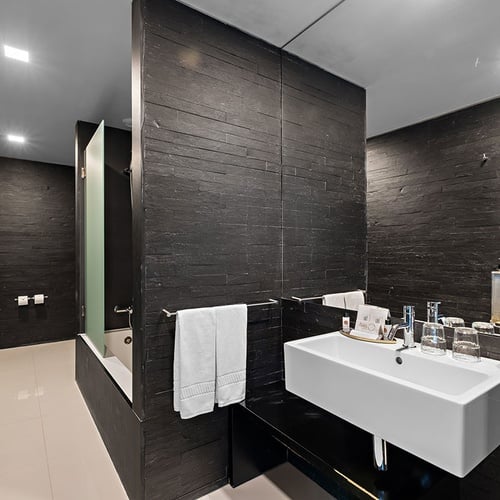 Um banheiro de hotel de design moderno, com paredes texturizadas de ardósia preta, piso claro e acessórios brancos, é retratado nesta imagem.