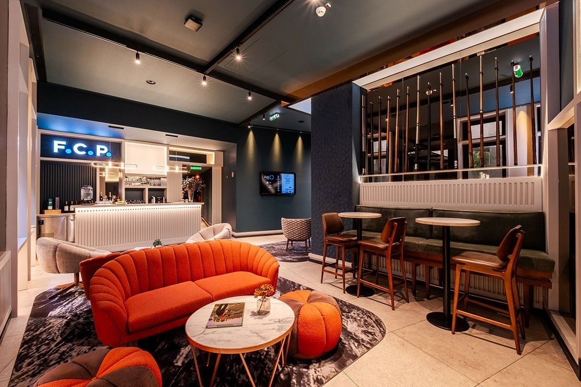 A imagem mostra uma área de bar/lounge moderna e elegante, com um sofá laranja vibrante, bancos de bar e um balcão iluminado com a inscrição 