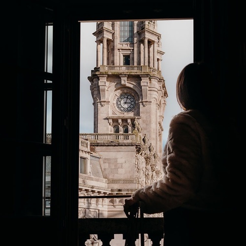 A person is silhouetted as they look out from a dark interior through an open window or balcony towards a grand architectural clock tower.