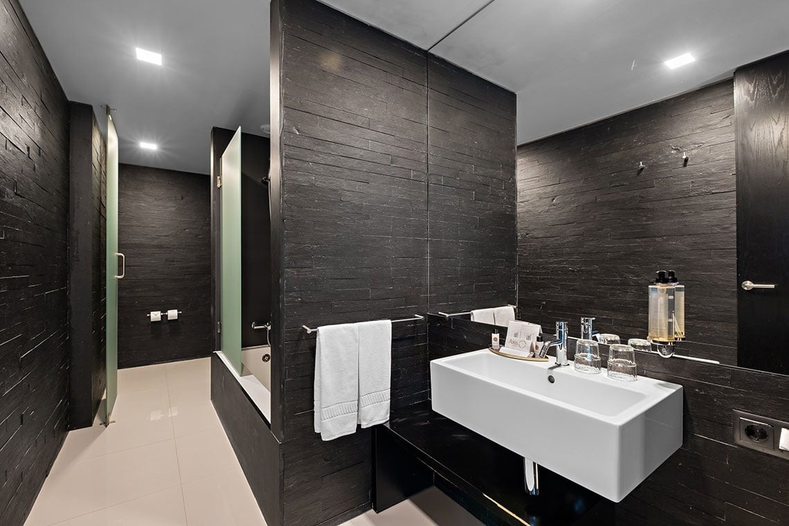 Une salle de bain moderne présente des murs texturés en ardoise noire, un lavabo blanc sur un comptoir sombre, et une zone de douche/baignoire partiellement fermée par des parois en verre dépoli.