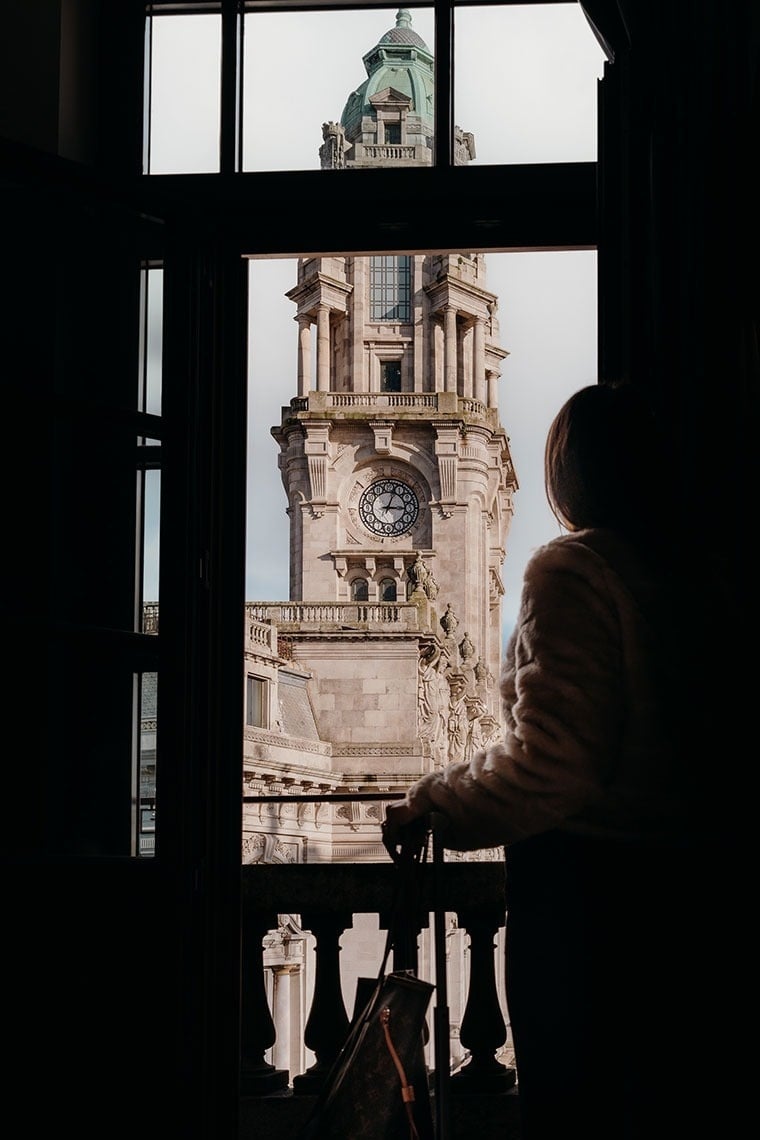 Uma silhueta de uma pessoa observa uma majestosa torre de relógio de pedra através de uma janela.