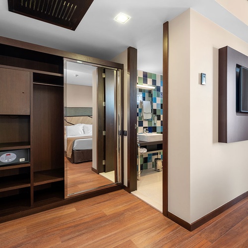 Um quarto de hotel moderno apresenta um guarda-roupa de madeira escura com espelho que reflete um quarto adjacente, uma entrada para um banheiro com azulejos coloridos e uma televisão de tela plana montada na parede.
