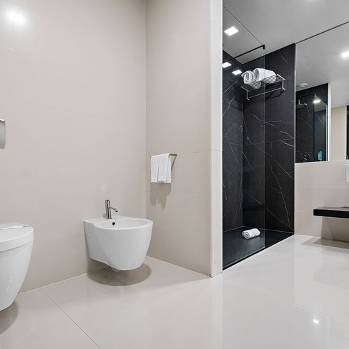 Um banheiro moderno e elegante com revestimentos claros, destacando um vaso sanitário, bidê, box de chuveiro com mármore escuro e pia suspensa.
