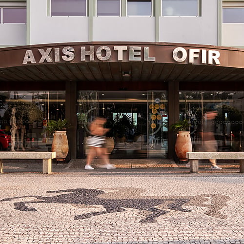 Entrada del Axis: Corporate Axis Hotel Ofir con personas borrosas en el exterior y un coche rojo reflejado en el cristal.