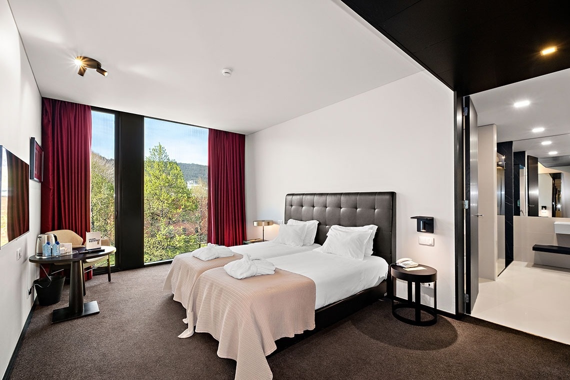 Cette photo représente une chambre d'hôtel moderne dotée de deux lits jumeaux, d'une grande fenêtre avec vue sur la nature et d'une salle de bain adjacente.