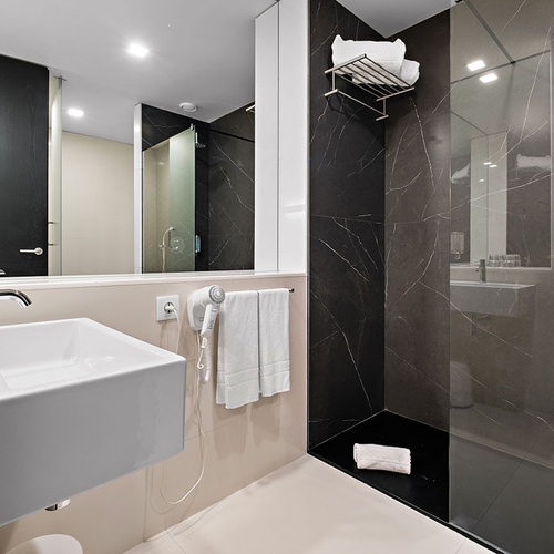 Uma casa de banho moderna com um lavatório branco suspenso, um espelho grande e uma área de duche revestida em mármore escuro.