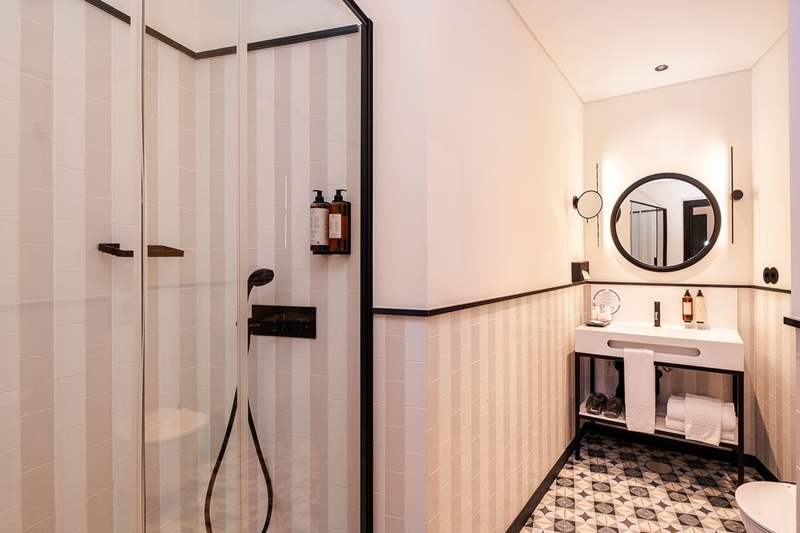 Um banheiro moderno apresenta um chuveiro com box de vidro e detalhes em preto, paredes com azulejos listrados em tons claros, e uma bancada com pia e espelho redondo iluminado, complementados por um piso com padrão geométrico.