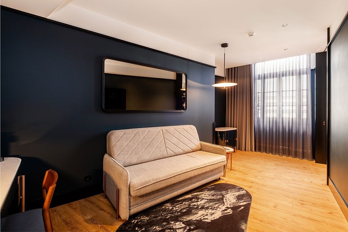 Um quarto moderno apresenta uma parede azul-marinho com um espelho retangular, um sofá-cama bege, piso de madeira claro e uma janela ampla com cortinas.