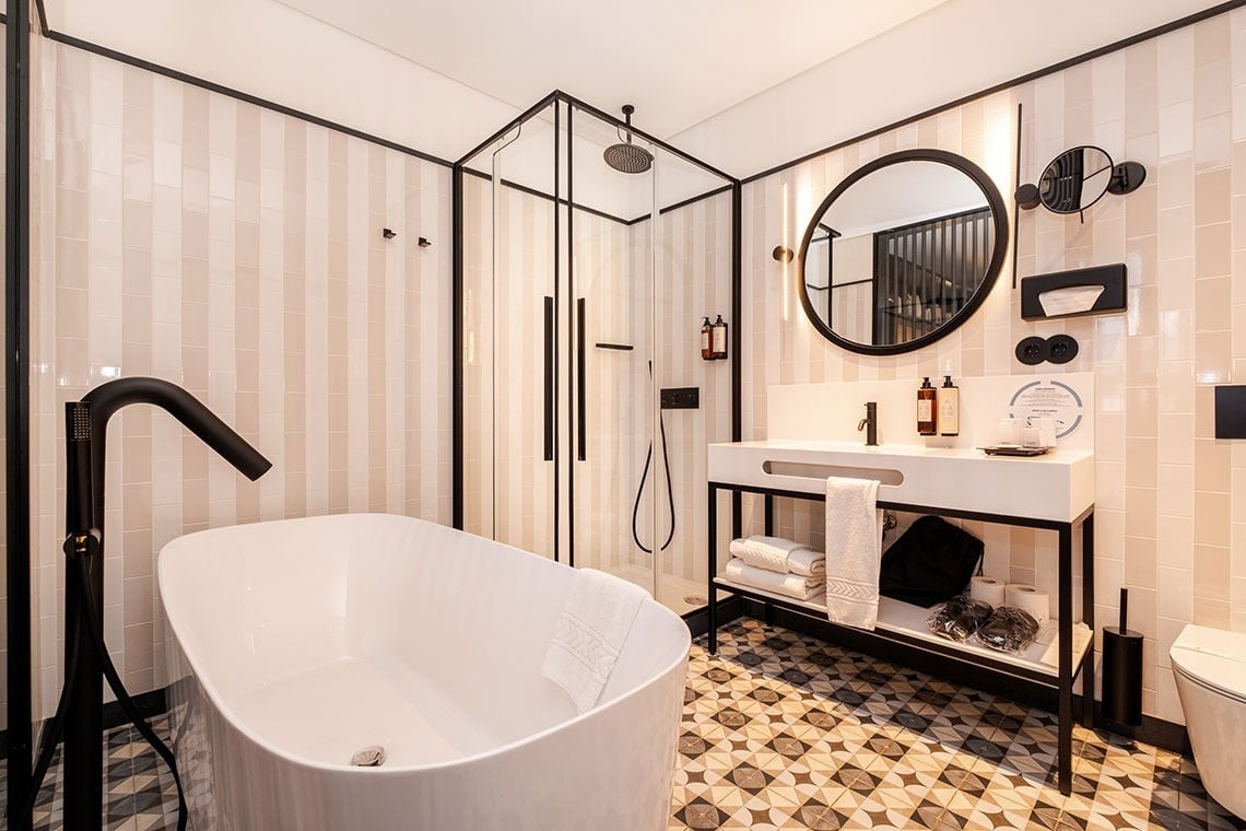 Um banheiro moderno e elegante com banheira e box de chuveiro de vidro, destacando-se pelos acabamentos pretos contrastantes e o piso geométrico estampado.