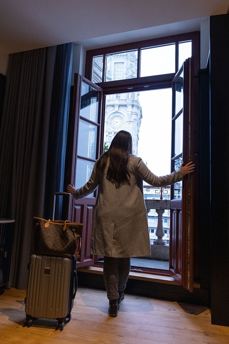 une femme avec une valise et un sac louis vuitton se tient devant une fenêtre ouverte
