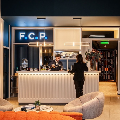 In a modern and stylish reception area or bar, a man pours wine for a woman standing at the counter, under a glowing 