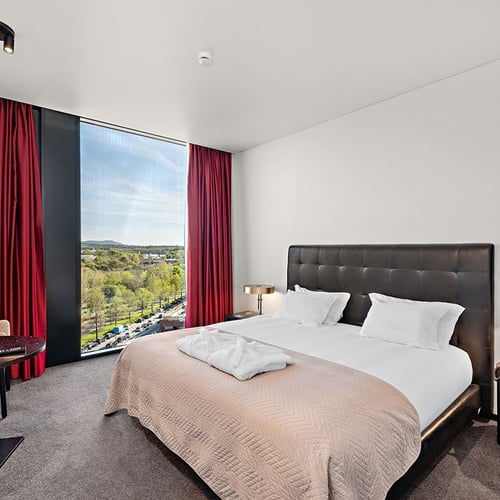 Um quarto de hotel moderno e luminoso apresenta uma cama de casal com cabeceira escura, cortinas vermelhas vibrantes ladeando uma grande janela com vista para uma paisagem verdejante e uma estrada.
