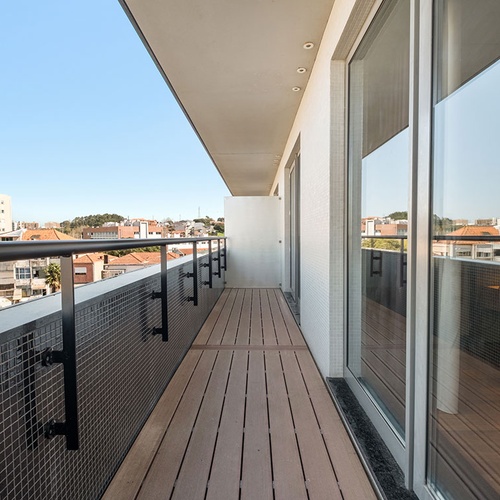 Uma varanda moderna e comprida, com piso de madeira e corrimão metálico sobre um muro de mosaico escuro, oferece uma vista urbana sob um céu azul, com grandes portas de vidro que refletem o ambiente.